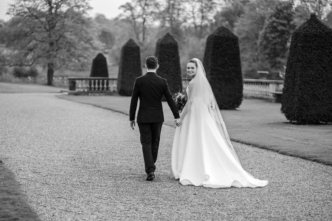 Black and white photograph of newlyweds walking down a tree-lined drive