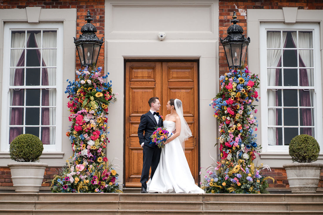 Bride and groom on the steps of a stately home framed by colourful flowers